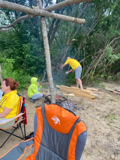 The image depicts an outdoor scene, possibly a picnic or barbecue, set against a backdrop of dense trees and vegetation. The ground is sandy, with scattered dry grass and twigs. In the foreground, two camping chairs are visible; one is occupied by a woman with red hair wearing a yellow t-shirt, and the other is empty. Further back, a person in a bright green hooded jacket is crouched down near a makeshift barbecue grill, attending to something on the grill. To the right, a man in a yellow t-shirt and gray shorts is standing and appears to be working with a stack of wooden planks. A rustic wooden structure, likely a shelter or support, is present in the middle ground. Smoke is rising from the grill, indicating cooking is in progress. The overall impression is a casual outdoor gathering in a natural, possibly wooded, area. The city of Ryabinino, Russia is not explicitly depicted or referenced in the visual information provided.