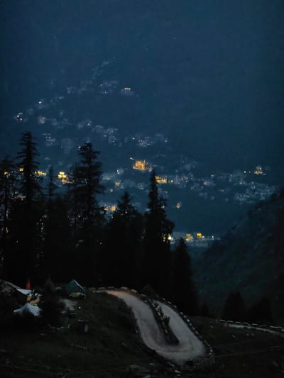 The image depicts a mountainous landscape at dusk or night, likely in or near Aleo, India, given the context. The foreground features a winding dirt road with some gravel and a few sparse streetlights or illuminated markers lining its edge. On the left side of the road, there are a few structures, including what appears to be a tent or camping shelter, and possibly a small building. The middle ground is dominated by dark silhouettes of pine trees, which are dense and cover a hillside. In the background, a city or town is visible spread across a valley and hillside. Numerous small lights from buildings illuminate the urban area, giving it a scattered, twinkling appearance against the dark mountainside. The overall atmosphere is dark and serene, with the distant city lights providing the only significant illumination besides the road.
