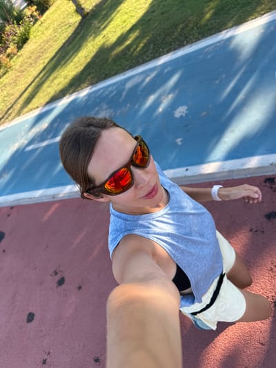 A woman is taking a selfie outdoors in Fethiye, Turkey, on a running track with a blue lane and a red lane. She is wearing a light blue tank top, white shorts with a black stripe, and sunglasses with orange mirrored lenses. Her hair is pulled back, and she has a white wristband on her left arm. Shadows of trees are cast across the track.