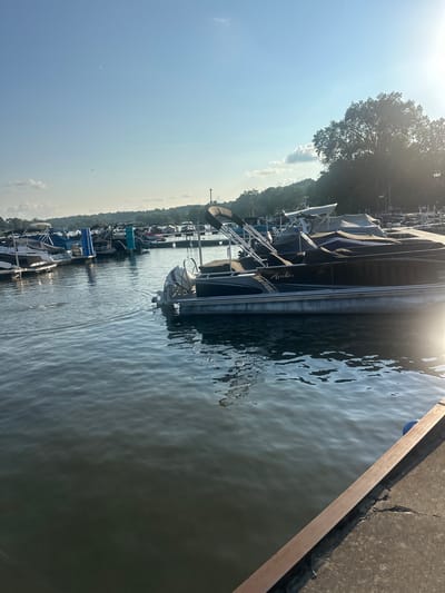 The image shows a marina on a sunny day. Several boats are docked on the left side of the frame, while a pontoon boat with the name "Avalon" visible on its side is prominently featured in the foreground on the right. The water is calm with reflections of the boats and the sky. Trees and a shoreline are visible in the background under a bright blue sky with some clouds and the sun shining. The location is the Fox River in Algonquin, Illinois, United States of America.