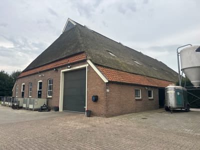 The image depicts a large, red brick barn with a thatched roof, situated in the city of Wilhelminaoord, Netherlands. The barn has a substantial roll-up garage door on its right side and several windows along its left facade. In front of the barn, a paved area is visible, with various objects and structures. To the left, there are several metal enclosures, possibly for livestock, and a pallet with equipment. To the right of the barn, a silver trailer with green doors is parked next to a large white silo. The sky is overcast with scattered clouds. The scene appears to be an agricultural or farm setting.