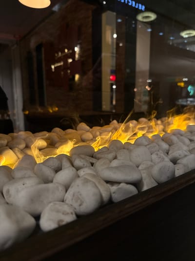 The image displays a low-angle view of an artificial fireplace within an indoor commercial setting in Jessheim, Norway. The primary subject is a dark-rimmed trough filled with numerous smooth, irregularly shaped white stones, from which yellow-orange flames and heat haze emanate. The flames are concentrated across the middle and right sections of the stone bed. A clear glass barrier separates the fireplace from the background, displaying significant reflections. These reflections include blurred bright lights, dark structural elements of a modern building, a distinct red light, and a mirrored sign fragment visible at the top, appearing to read "REFLECKED." No discernible individuals are present within the frame. The background visible through the glass is dark and indistinct, suggesting a night scene or dimly lit area outside or adjacent to the interior space.