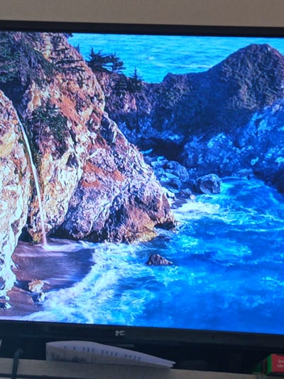 This is a low-angle, blurry photograph of a television screen displaying a scenic view of a rocky coastline with a waterfall and turquoise water. The image on the screen features a dramatic cliff face with jagged rocks, some covered in greenery, and a thin waterfall cascading down onto a small beach below. The ocean is a vibrant blue, with white foam indicating waves breaking near the shore. In the background, more rocky formations rise out of the water. The bottom of the television screen is visible, showing a black bezel and a small, white object with some text on it placed on a surface in front of the screen. The scene is not in Litomyšl, Czechia.