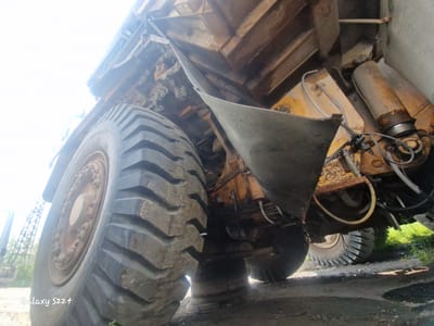 The image shows a low-angle, close-up view of the underside of a large, industrial dump truck, likely parked outdoors in Zheleznogorsk-Ilimsky, Russia. The massive tire of the truck dominates the left side of the frame, with visible tread patterns and some dirt accumulation. The truck's undercarriage, including hydraulic cylinders, metal bracing, and yellow painted components, is visible. A curved metal shield hangs down from the truck's body. In the background, a portion of a tower structure and some greenery can be seen. The overall impression is one of heavy machinery in an industrial setting. The watermark "Galaxy S22+" is visible in the lower left corner.