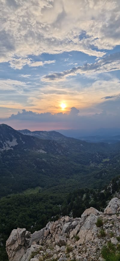 An aerial view captures a sunset over a mountainous landscape. The sun, a bright orb with visible rays, is positioned centrally in the sky, casting a warm orange and yellow glow that blends into softer pinks and purples towards the horizon. Clouds, varied in texture and density, drift across the sky, some catching the sunlight and appearing illuminated.

Below the sky, the terrain transitions into a series of rugged mountains. The closest mountains in the foreground are rocky and arid, with sharp, angular rock formations. These give way to a dense forest that carpets the slopes and valleys of the mid-ground and background. The trees are a deep, rich green, suggesting a thick, verdant growth. Further in the distance, successive mountain ranges recede into a hazy, muted blue, indicating atmospheric perspective. The overall impression is one of a remote, natural environment bathed in the soft light of dusk. The location is identified as Ubla, Bosnia and Herzegovina.