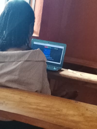 A person with braided hair, wearing a striped shirt, is seated at a wooden bench and using a laptop computer. The laptop is on a wooden surface. The background shows a dark red wall. The location is Oke Ogun, Nigeria.