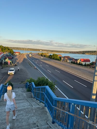 A blonde woman in a white t-shirt and denim shorts walks down concrete stairs with a blue railing. She is facing away from the camera. The stairs are part of a pedestrian overpass. Below the overpass is a multi-lane highway with cars and trucks. On the left side of the highway are several small buildings and a white car. On the right side of the highway, there are houses and trees. A large body of water, likely a river or a reservoir, is visible in the distance, with hills on the far side. The sky is clear and blue with some light clouds. The sun appears to be low in the sky, casting long shadows. The overall scene depicts a roadside landscape in Yelyzavetivka, Ukraine.