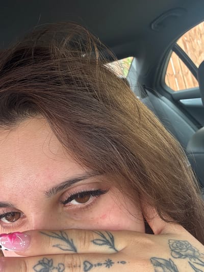 The image shows a close-up of a person's face, with their hand covering their mouth. The person has brown hair and brown eyes, and is wearing eyeliner and false eyelashes. Their fingernails are decorated with pink and white patterns. There are tattoos visible on their hand. The background suggests the person is inside a car, with a view of a wooden fence through the car window. The location of Dungannon, United Kingdom, is not discernible from the image itself.
