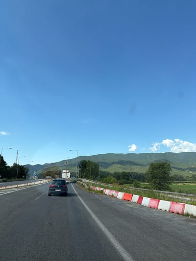 This image depicts a highway scene in the Municipal Unit of Promachonas, Greece, under a clear blue sky with scattered white clouds. The road is asphalt with white lane markings. A dark-colored Volkswagen Golf hatchback is driving in the right lane, facing away from the viewer, with its license plate reading "BP 973 CB". To the right of the road, there is a barrier made of alternating red and white plastic traffic cones and bollards. Further ahead, a white truck with "TRANSPORT" written on its side is visible, along with traffic lights displaying amber. To the left of the road, there are orange traffic cones and signage. In the background, lush green mountains are visible, with trees and vegetation in the foreground and midground. The overall scene suggests a typical daytime view of a road in a rural or semi-rural mountainous area.