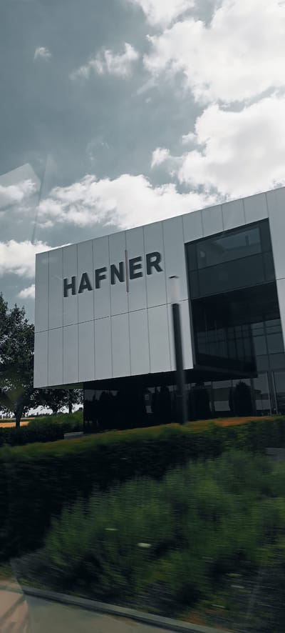 The image shows the facade of the "HAFNER" building, a modern commercial structure with a white, panel-covered exterior. The building features large, dark-tinted windows and a prominent sign with the company name. It is situated in Fellbach, Germany, where it serves as a business establishment. The foreground shows a blur of green foliage, suggesting the photograph was taken from a moving vehicle. The sky above is overcast with a mix of light and dark clouds.