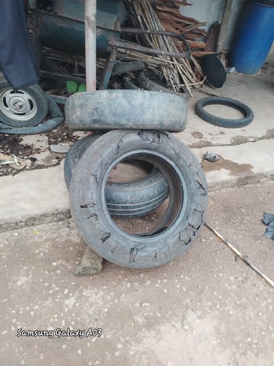 In Dokan Tofa, Nigeria, an outdoor scene depicts several discarded tires. In the foreground, a heavily damaged tire lies on the ground, showing significant wear and tear with several large holes and cracked rubber. Another tire is stacked on top of it, and a third tire is positioned behind these, partially obscured. To the left, a flat tire with a silver rim is visible, and beyond that, a dark blue fabric is draped over something unseen. The background reveals a cluttered area with wooden debris, metal structures, and a large blue plastic barrel. The ground is a mixture of concrete and dirt, with scattered stones and some damp patches. A thin stick lies horizontally across the lower right portion of the image. The overall impression is one of a disused or storage area for scrap materials.