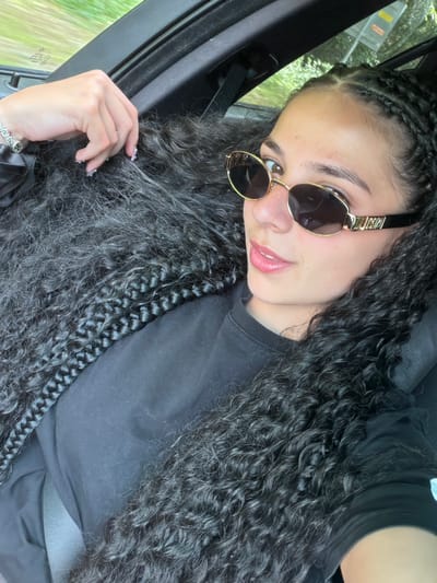 The media file shows a young woman with braids and long, curly dark hair, wearing sunglasses and a black t-shirt. She is taking a selfie while seated in a car. The woman has on-trend sunglasses with gold frames and dark lenses. Her makeup appears to include lip gloss. Her nails are decorated with small white accents. She is wearing a silver bracelet on her left wrist. The setting is inside a car, with the interior visible along the left side of the frame, including a dark car door and what appears to be a window showing blurred green foliage outside, suggesting movement. The lighting indicates it is daytime, and the scene has a casual, personal feel. The woman's facial expression is relaxed and she appears to be posing for the photo.