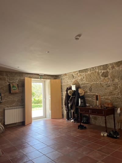 The image displays an interior room, likely an entryway, within a building located in Oliveira de Frades, Portugal. The room features robust walls constructed from irregular, light brown and grey exposed stone masonry. The ceiling is flat, white, and equipped with recessed circular spotlights and a centrally mounted white smoke detector. A green and white emergency exit sign is fixed above a wide, central doorway at the far end of the room. This doorway is composed of a white-framed glass door, open to an exterior view of bright green foliage under sunlight, and flanked by two tall, light-colored wooden panels. The floor is covered with square, reddish-brown terracotta-style tiles.

Along the left wall, a white horizontal panel radiator is installed. Above it, a brown paper bag, partially visible and displaying the text "FRESIA," hangs from the stone wall. On the right side of the room, against the stone wall, stands a dark wooden console table with turned legs and two drawers. Various items are placed on its surface, including a wooden box containing a wine bottle, a small wicker basket, and several small miscellaneous objects. Behind the table, a coat rack is laden with multiple dark jackets and coats, two hats (one straw-brimmed), and a dark bag with "LER" and "turismo rural" text. On the floor in front of and to the right of the console table, several pairs of footwear are present, including dark boots, black athletic shoes, and light-colored sneakers. A portion of a light-colored wooden chair is visible on the far right edge of the frame. The room is illuminated primarily by natural light emanating from the open doorway. No individuals are present in the scene.