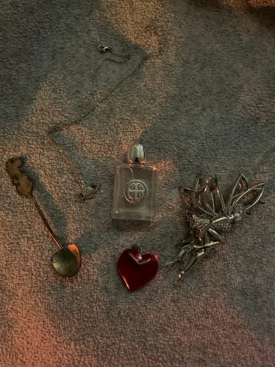 A collection of five distinct objects is displayed on a textured, grey fabric surface within a dimly lit environment, illuminated by a warm light source from the lower-left, in Choctaw, United States. No individuals are present. The objects are arranged across the surface: In the upper-left, a silver-colored chain necklace with a small, orb-shaped, stone-set pendant. To its lower-left, a small, dark-patinated metal spoon with a decorative handle and a heart-shaped bowl. Centrally, a clear, rectangular glass bottle with a silver cap, featuring a circular St. Benedict Medal embedded on its face. Below the bottle, a translucent red heart-shaped pendant. To the right of the bottle, a detailed, silver-colored metal figurine or brooch depicting a winged fairy.