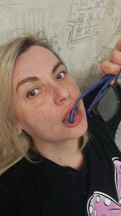 An indoor scene in Bel'kovo, Russia, features a blonde-haired woman with fair skin, brown eyes, and dark eyebrows, directly facing the camera with a wide-eyed expression. She wears a black v-neck shirt featuring a pink and white graphic on the lower right. Her right hand holds a pair of blue-framed sunglasses with dark lenses, and one arm of the sunglasses is positioned inside her mouth, between her lips. The background is a light-colored wallpaper patterned with monochrome line drawings of architectural structures, including multi-story buildings and domes.