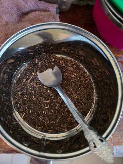 A close-up, top-down view shows a metal pot filled with loose-leaf tea. A silver spoon rests in the tea, with its handle extending out of the pot. The pot is on a wooden surface. In the background, a pink towel and part of a pink bowl are visible. The scene appears to be indoors.