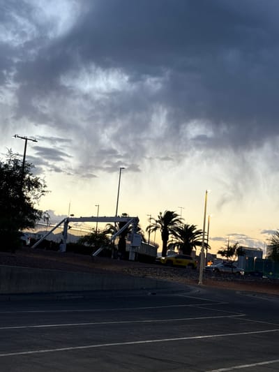 The image depicts an outdoor scene in Paradise, Nevada, United States. The sky is predominantly filled with a mix of dark, ominous clouds and lighter, brighter areas indicating the presence of sunlight or an approaching sunset. Silhouetted palm trees are visible, indicating a warm climate or tropical theme. A chain-link fence and light poles are present. A section of paved road or parking lot with painted lines is visible on the right, suggesting the image was taken from a street-level perspective.
