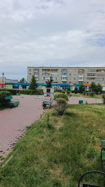 The image depicts a public space with a monument, benches, and a residential building in the background. The monument, centrally located, features two figures, one appearing to be an adult holding a child. A wreath of flowers is placed at its base. Two green park benches are visible in the foreground, one on the left and another on the right, partially obscured by a bush. The ground is paved with red bricks in a patterned walkway, and a grassy area with some dry patches borders it. The residential building behind the monument is a multi-story, light-colored structure with numerous windows and balconies. In front of the building, a strip of shops with colorful signage is visible. The sky is overcast with clouds, suggesting a cloudy day, and no specific time of day can be determined. The overall scene appears to be a quiet, everyday moment in a town square or park. The setting is identified as Barabinsk, Russia. There is some visible text on the shop signs, but it is not clearly legible.