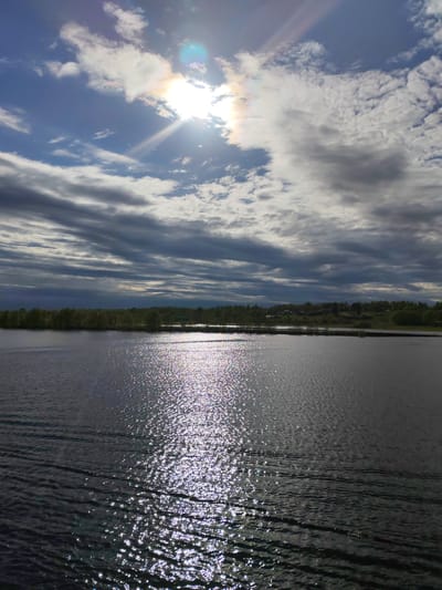 The image shows a body of water under a sky filled with clouds. The sun is visible, obscured by some clouds, creating a bright glare and radiating light rays in the sky. The water's surface reflects the sunlight, causing a shimmering effect. Across the water, a strip of land with trees is visible. The photo was taken in the city of Zelonaya Roshcha, Russia. No people are visible.