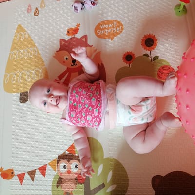A bald baby with blue eyes and an open mouth lies on its back on a playmat. The baby wears a pink top with a strawberry print and a diaper. The playmat is white and features colorful cartoon graphics of animals and natural elements, including a fox, an owl, a bird, trees, and sunflowers. To the baby's right, a large pink spiked ball is partially visible. The scene is lit by natural light. The image is oriented with the baby's head towards the bottom left.