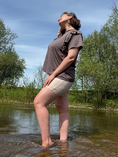 A medium shot shows a woman standing in a shallow body of water, possibly a river or stream, in Усть-Тунтор, Russia. The woman is wearing a short-sleeved, gray-brown shirt and light-colored shorts. She is looking towards the water and is in a slightly bent-over position. The water line reaches around her calves. The background shows green foliage, suggesting a wooded or grassy area. Blue sky is visible above, partly obscured by tree foliage.