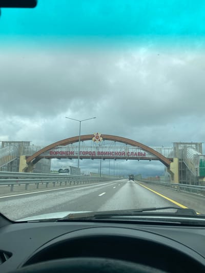 A view from inside a vehicle, looking toward an arched sign on a roadway. The sign features the text "Voronezh - City of Military Glory" in Russian. The roadway is a multi-lane highway with yellow lines. The weather is overcast, and the surroundings consist of industrial infrastructure, potentially on the outskirts of Svkh Voronezhskiy, Russia. Other vehicles are visible on the highway.
