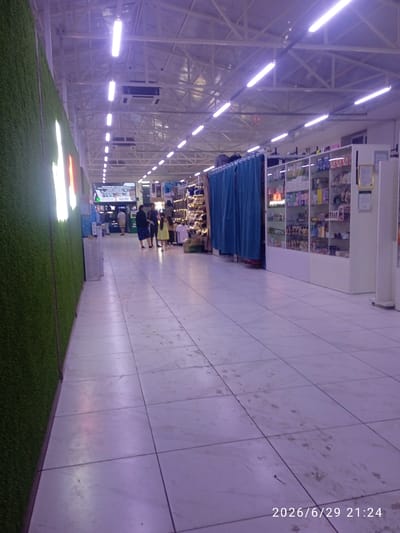 The image shows the interior of a market or retail store in Ak-Suu, Kyrgyzstan. The scene is illuminated by fluorescent lighting mounted on the high, white, corrugated metal ceiling. The floor is tiled in a light color, with a visible grid pattern and some scuff marks. To the left, a long wall is covered with artificial green turf, with a vertical, illuminated sign.
In the mid-ground, several individuals are visible. A family group, including at least two adults and a child in a yellow dress, are walking towards the right side of the image. Further down the aisle, other shoppers are present, some browsing merchandise. On the right side of the image, there are shelves stocked with various products, seemingly in a retail display. Blue curtains are visible, likely separating sections of the store. A large digital display screen is mounted on a wall in the background. The overall atmosphere appears to be that of a busy commercial space. The date and time "2026/6/29 21:24" is superimposed on the bottom right corner of the image.