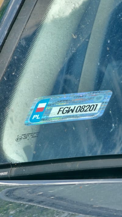 A zoomed-in, eye-level shot focuses on a windshield sticker. The sticker is rectangular with a holographic, shimmering blue background and a white stripe running horizontally across the center. On the left side of the sticker, there is a blue rectangle with the Polish flag (red and white) in the top right corner, and "PL" in white text below it. To the right of this, the white stripe on the sticker contains black text that reads "FGW 08201". Below the sticker, etched into the glass, are smaller markings including "SAINT-GOBAIN SEKURIT", "E20", "43R", and "00348". The windshield is dusty, with a few small specks of debris visible. The background outside the windshield is blurred and dark. The provided metadata indicates the image was taken in Hristo Danovo, Bulgaria.