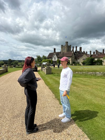 The image captures a frontal view of a large, stately manor house with a grey, overcast sky. In the foreground, a gravel driveway leads towards the house. On the left side of the driveway, a woman with long, dark red hair, wearing a black jacket and black pants, faces to her right, holding a small, light-colored object in her hand. To the right of the driveway and slightly in front of the woman, a man wearing a white long-sleeved shirt, blue jeans, and a pink baseball cap faces to his left. He is holding a bright yellow banana in his right hand. Behind the couple, the manor house is a prominent structure with multiple chimneys, various architectural elements, and a flag flying from a tower. The grounds surrounding the house consist of well-maintained green lawns and some stone structures. The scene appears to be a tourist or visitor location, possibly a historic estate. The image does not provide any explicit visual cues to confirm the location as being within the city of Chichester, United Kingdom.