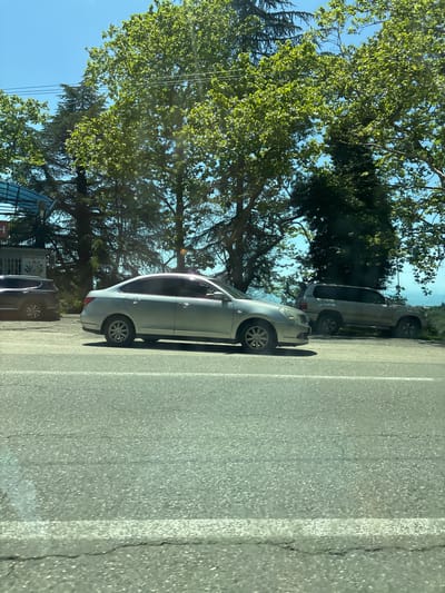 The image, taken in Viadati Eshyra, Abkhazia, depicts a sunny daytime scene along a paved road. The perspective appears to be from a vehicle, looking towards the right side of the road, which is flanked by trees and parked cars.
In the mid-ground, a light-colored (likely silver or light grey) sedan is parked parallel to the road. Behind it, further back and to the right, a white SUV is also parked. A dark-colored car is partially visible in the bottom left, parked near a building.
The upper and left parts of the scene are filled with a variety of trees, including lush green deciduous trees with bright foliage and darker coniferous trees, suggesting a verdant environment. Thin wires or cables are seen extending downwards from the trees in the upper left. In the distant background, beyond the white SUV, a body of water—possibly a lake or the sea—is visible under a clear blue sky.
The road surface is asphalt, with distinct white lines marking its edge and possibly a parking zone. Bright sunlight creates visible shadows and reflections on the road and vehicles.
On the far left, behind the dark-colored car, a small building or kiosk with a blue awning is visible. The building features Russian text that reads "ПРОДУКТЫ" (PRODUKTY), which translates to "GROCERIES" or "PRODUCTS," indicating a local store. No people are visible in the image. The scene conveys a tranquil, sunny day in a natural setting adjacent to a roadway.