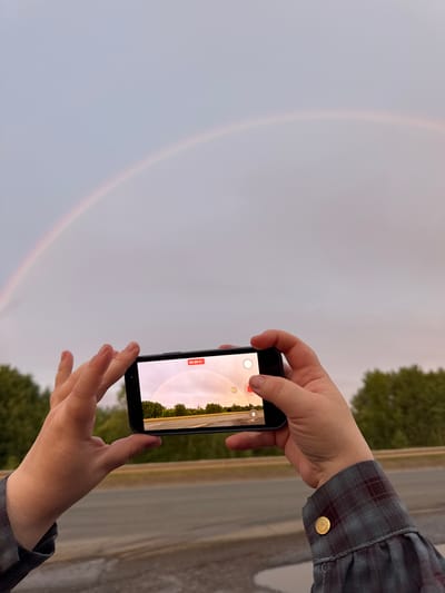The media file is a photograph taken outdoors, likely in Sovkhoznyi, Russia. The image shows a person's hands holding a smartphone, capturing a faint rainbow in a cloudy sky. The smartphone screen displays a video recording interface with a red record button and a timer showing "00:00:01". Green trees and a paved road are visible in the background, suggesting a rural or suburban setting. The weather appears overcast, with the rainbow indicating recent rain or atmospheric moisture. The person is wearing a plaid shirt with a brass button on the cuff, and their hands are positioned to hold the phone steady. The overall mood is one of observation and appreciation of a natural phenomenon.