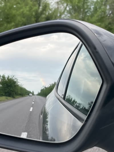 The image shows the reflection in a car's side mirror. The reflection includes a road with white lane markings, green vegetation on either side, a car in the distance, and a cloudy sky with what appears to be a rainbow. The location is unknown and not determinable from the image. There is no indication of Sofiivka, Ukraine.