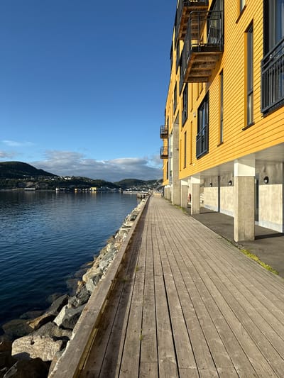 A waterfront promenade in Harstad, Norway, runs alongside a bright yellow building with balconies. The promenade is constructed of weathered wooden planks and is bordered by a rocky embankment. The water, a deep blue, gently laps against the rocks. In the background, a hillside town with houses and buildings is visible under a clear blue sky with a few scattered clouds. The scene is devoid of any people.