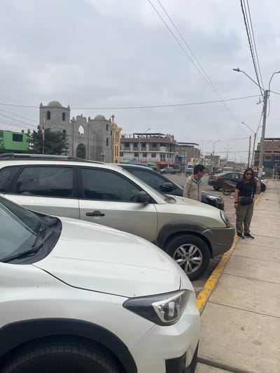 The scene is set in Quilmaná, Peru, on an overcast day. In the foreground, two parked cars dominate the view: a white car with a black fender and a gray car slightly behind it.

Two individuals are visible on the sidewalk adjacent to the cars. One is a woman with dark hair and sunglasses, dressed in a black top and green pants. She stands with her hands in front of her. Next to her is a man in a tan jacket and glasses, standing with his head tilted.

Behind the parked cars, a cityscape emerges, featuring buildings of various heights and colors. The most prominent structure is a large, unfinished church or religious building with arches, domes, and a cross on top. Utility poles with wires traverse the sky. The sidewalk is painted yellow and gray.