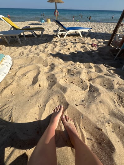 The image shows a beach scene, likely in Mrezga, Tunisia. The main subject is a patch of sandy beach with a person's feet visible on the right side. The person is standing, with their legs casting a shadow on the sand. The sand is light brown in color, with scattered pieces of debris. In the background, there are beach chairs, and what appears to be a life preserver attached to a pole. Further back, the blue sea is visible with people swimming and floating in the water. The sky is clear and blue.