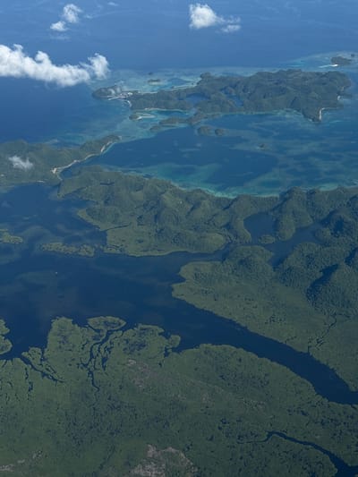 This is an aerial view of a coastal landscape, likely in a tropical or subtropical region. The scene features a mix of land and water. The land is heavily vegetated with dense green foliage, suggesting lush forest or mangrove ecosystems. There are numerous waterways, channels, and inlets cutting through the land. The water ranges in color from light turquoise to deep blue, potentially indicating varying depths or the presence of coral reefs or sandbars. In the foreground, there's a series of small islands or land formations surrounded by shallow, clear water, with visible coral reefs. Clouds are visible on the left side of the image.
Based on the request for the city of Del Carmen, Philippines, this image likely shows the Bucas Grande Island area near Del Carmen, Surigao del Norte. The area is characterized by its unique karstic limestone formations, lagoons, caves, and mangrove forests.