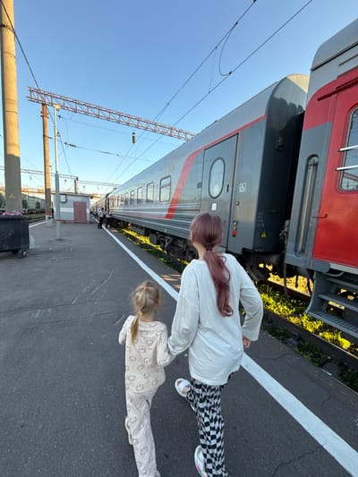 In Uzunovo, Russia, two young girls walk along a train platform beside a train. The girl in the foreground wears a light-colored pajama set with a bear print, while the girl in front of her has pink-tinged hair and wears a white long-sleeved shirt with black and white checkered pants. They are walking away from the camera, holding hands. The train, a passenger car with gray and red accents, is parked on the tracks. Overhead, power lines and supporting structures are visible against a clear blue sky. The platform is paved with asphalt, marked by a white line along the edge. A small utilitarian building and a trash bin are also present on the platform. The scene appears to be a train station.
