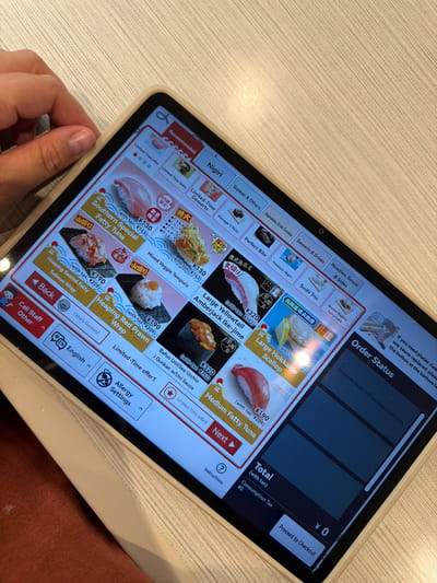 This is a close-up, eye-level, indoor shot of a person's hand holding a tablet computer, which is displaying a sushi restaurant's digital menu. The tablet is positioned on a light-colored wooden surface, possibly a table. The menu features various sushi items, with prices and descriptions, and the "Order Status" section on the right side of the screen indicates "Ordered items will be displayed here." The background is blurred, suggesting the focus is on the tablet and the user's interaction with it. The restaurant appears to be located in Chiyoda, Japan, as indicated by the contextual information.