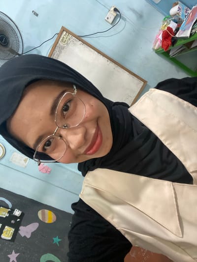 The image is a selfie taken in what appears to be a classroom or office setting. A young woman wearing a black hijab and glasses is smiling at the camera. She is wearing a black top and a cream-colored vest. In the background, there is a light blue wall, a fan, a whiteboard, and shelves with various items. Part of a dark surface with colorful drawings of space elements like planets and rockets is visible at the bottom of the image. The overall impression is of a casual, indoor environment. The location of Tanjung Gading, Indonesia is not evident from the visual elements present in the photograph.
