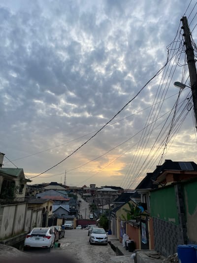 The image depicts a street scene in Isheri, Nigeria. Overcast skies with some golden light fill the majority of the frame. Utility poles and a dense network of electrical wires are visible at the top of the frame. The street is lined with buildings of varying heights and architectural styles, some featuring solar panels on their roofs. Parked cars line one side of the street. A person is walking on the right side of the road. The scene is taken from a low angle, looking upwards.