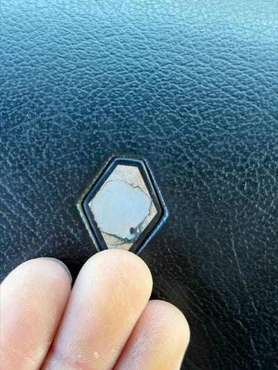 The image is a close-up, macro shot of a tarnished, hexagonal metal emblem embedded in a textured black surface, possibly leather or a similar material. The emblem has a light-colored, stone-like insert with visible cracks and discoloration, surrounded by a dark metallic frame. A portion of a person's fingers, showing distinct fingerprints and dry skin, are visible in the lower left corner, suggesting they are interacting with or pointing at the emblem. The background is out of focus, but shows a consistent, slightly rough texture. The lighting appears to be natural, casting subtle shadows and highlighting the details of the emblem and the texture of the surface. The scene is devoid of any specific urban or natural elements that would indicate a location in Zarech'e, Russia.