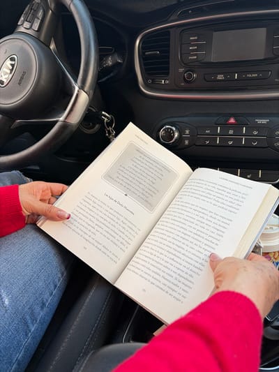 The image shows a person sitting in the driver's seat of a KIA car, holding open a book titled "La Dona Huevotes Y Los Hijos." The book appears to be in Spanish and is opened to pages 134 and 135. Page 134 references "Las Hijas de Dona Huevotes." Page 135 has text about how a woman's relationships with other women in her life impacts how she sees herself. The person is wearing a red top and blue jeans. The car dashboard and steering wheel are also visible. The car keys are dangling from the steering column.