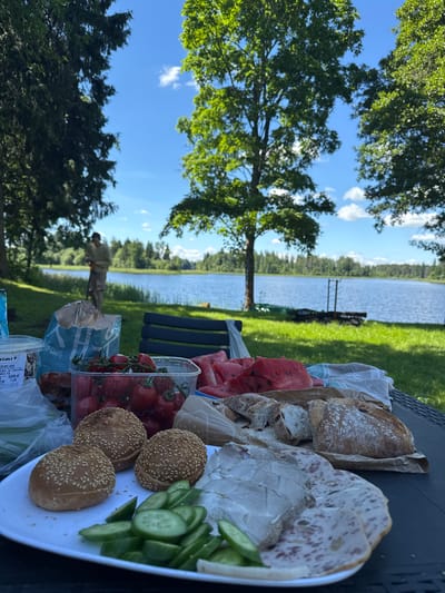This image depicts a picnic spread on a table near a lake in Lendži, Latvia. In the foreground, a black table holds a variety of food items: a white plate with three sesame seed buns and sliced cucumbers, a plastic container overflowing with cherry tomatoes, slices of watermelon, a partially unwrapped loaf of bread, and several types of sliced deli meats. A blue bag with "CHIPS" printed on it is visible. In the background, a person in a light-colored shirt and shorts stands near the water's edge, facing away from the camera. The lake stretches across the mid-ground, bordered by lush green grass and trees. The sky above is blue with scattered white clouds, and the overall scene suggests a sunny day.