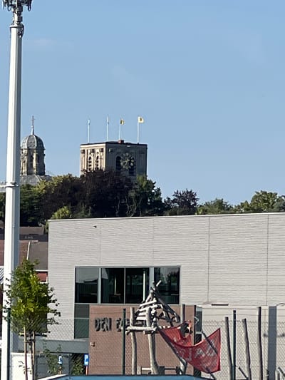 This is a vertical, outdoor, daytime photograph of a street scene in Scherpenheuvel, Belgium. The image is dominated by a modern, grey, corrugated building on the right side of the frame. This building has several large windows, some reflecting the sky. To the left of this building, a section of a dark brick building is visible, with some text that appears to be in a non-Latin script. In front of the brick building, there is an abstract sculpture made of light-colored wood, topped with a red banner that has white text on it.

Further to the left, a tall clock tower rises, adorned with architectural details and flying three small yellow flags. Below the clock tower, the dome of another building can be seen peeking out from behind trees. Several trees with green and dark foliage are interspersed throughout the scene, contributing to the overall urban setting.

In the foreground, a large white beam runs horizontally across the bottom of the image, suggesting the presence of a walkway or part of a structure. The sky is clear and blue, indicating fair weather. The overall impression is one of a bright, sunny day in a European town with a mix of historical and modern architecture. No people are clearly visible in the image, and the focus is on the buildings and architectural elements.