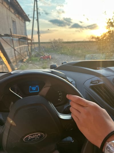 A person's hand is visible on the steering wheel of a Ford vehicle, as seen from the driver's perspective inside the car. The steering wheel has a visible "Ford" logo and "AIRBAG" marking. The car's dashboard displays a temperature of 31°C and the message "Vehicle is on." Outside the car, a rural scene is visible through the windshield, featuring a building with scaffolding, utility poles, a field, and a bright sunset with sun flares. A small red toy vehicle is on the ground near the building. The scene appears to be in Mlekarevo, Bulgaria, as indicated by the presence of a rural landscape with typical Bulgarian infrastructure.