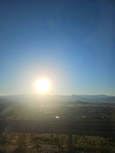 The image captures a sun-drenched vista from a slightly elevated vantage point overlooking a town nestled amongst rolling hills and distant mountains. The sky is a clear, bright blue, with the sun positioned in the upper left quadrant, casting a brilliant glow that creates lens flare and washes out details in its immediate vicinity. A faint, smaller circular anomaly is visible in the sky to the right of the sun.

In the mid-ground, the town of Gornji Kokoti, Montenegro, is spread out, characterized by a dense collection of buildings and structures, interspersed with patches of greenery. Some agricultural fields or undeveloped land are visible surrounding the town. The light suggests it is likely morning or late afternoon, with the low angle of the sun creating long shadows and a hazy, golden atmosphere.

In the foreground, a dark, weathered guardrail runs horizontally across the lower portion of the image, indicating the viewpoint is likely from a road or elevated path. The ground in front of the guardrail is covered in dry, sparse vegetation, including rocks and what appear to be dry grasses and some scrub brush. The overall impression is one of a rural landscape bathed in the warm light of the sun. There are no people or animals visible in the image.