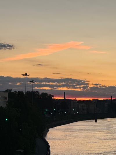 This image depicts a sunset over the city of Ladozhskii Trudposelok, Russia, with a river in the foreground. The sky is painted with hues of orange, pink, and pale yellow, with streaks of clouds running horizontally. In the distance, a silhouette of buildings and industrial chimneys can be seen against the colorful sky. A river reflects the sunset colors, with a small dark object, possibly a buoy or a submerged structure, visible in the water. The left side of the image shows a dark, tree-lined embankment leading to a pathway. Tall lampposts stand silhouetted against the sky. Green lights are visible along the embankment, suggesting streetlights or traffic signals. The overall scene is serene and atmospheric, capturing the quiet beauty of an evening in the city.