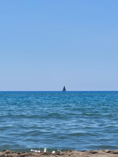 The image depicts a serene seascape at Castiglione della Pescaia, Italy. The horizon line divides the frame into two distinct halves: the upper half is a clear, vast expanse of light blue sky, devoid of any clouds. The lower half is dominated by the deep blue of the sea, with gentle waves reflecting the sunlight, creating a shimmering effect on the surface. In the distance, a lone sailboat with a dark sail navigates the water, appearing as a small, solitary silhouette against the expansive seascape. The foreground shows a strip of sandy beach, slightly out of focus, with some small, sunlit highlights on the sand near the water's edge. The overall impression is one of tranquility and a calm, sunny day at the coast.