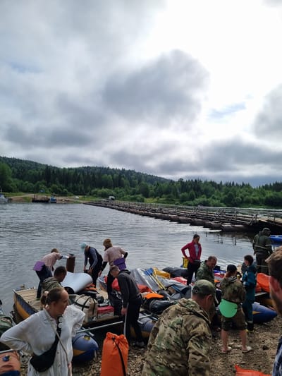 The image depicts a group of people on a riverbank near a pontoon bridge. The scene appears to be in Vishegorsk, Russia, given the context. In the foreground, several individuals are gathered around inflatable rafts, some of which are loaded with gear. One woman in a white shirt is looking to the right, with a black crossbody bag. Another person in a camouflage jacket is walking away from the camera.
Further back, more people are engaged in activities related to the rafts, with some appearing to be preparing for a journey. A pontoon bridge, constructed from logs and supported by pontoons, stretches across the river in the mid-ground, leading to a forested shoreline on the opposite bank. The sky is overcast with dramatic clouds, and the water of the river is visible in the foreground and mid-ground, reflecting the sky. The shoreline is rocky and covered with gravel. Several boats are docked on the left side of the river. The overall impression is of an outdoor recreational or travel setting.