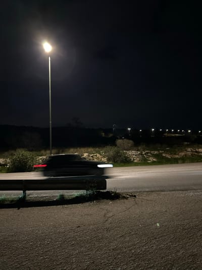 The image captures a nocturnal scene on what appears to be a rural or semi-rural road in Anatoliki Attiki, Greece. The photograph is taken from an elevated perspective, looking down and along the road.

In the foreground, a tall streetlight on the left emits a bright, white glow, illuminating the immediate surroundings and casting a strong lens flare. Parallel to the camera's perspective, a dark metal guardrail runs along the edge of the asphalt road. The road surface, which appears somewhat reflective, suggesting it might be wet or damp, extends into the distance.

In the midground, beyond the guardrail, a landscape of rocky terrain, sparse bushes, and small trees is visible, partially lit by the streetlights. A vehicle is discernible on the road, represented by blurred lights. A white, elongated blur indicates a headlight or front light, while a distinct red streak behind it signifies a taillight, suggesting a car moving away from the viewer or captured with a slight long exposure.

In the background, a long line of evenly spaced, smaller bright lights extends into the dark night, following the curve of the road into the horizon. These are likely more streetlights, providing illumination along the path. The sky above is profoundly dark, indicating the deep night. There is no visible text in the image. The overall atmosphere is quiet and solitary, characteristic of a road at night.